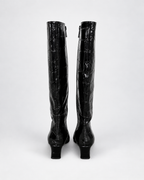 A pair of black knee-high boots with a glossy crocodile-embossed texture, shown from the back against a light gray studio background.