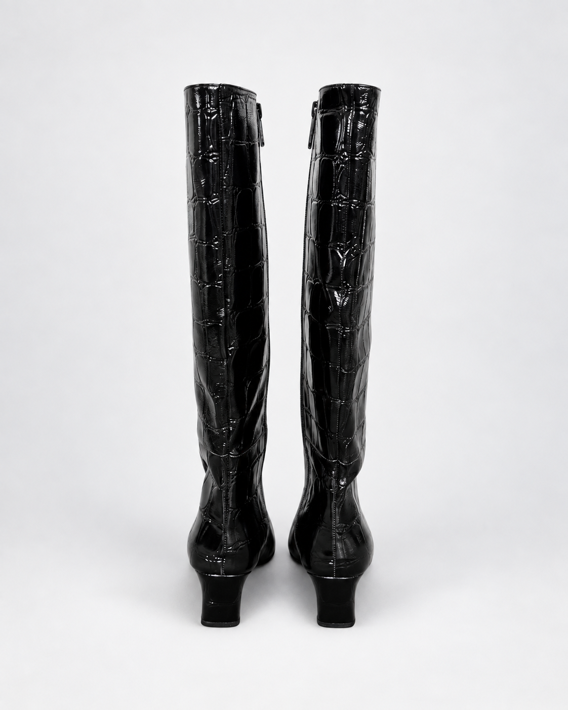 A pair of black knee-high boots with a glossy crocodile-embossed texture, shown from the back against a light gray studio background.